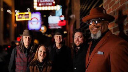 Five musicians pose on a city sidewalk at night, lit by colorful neon signs from nearby venues.