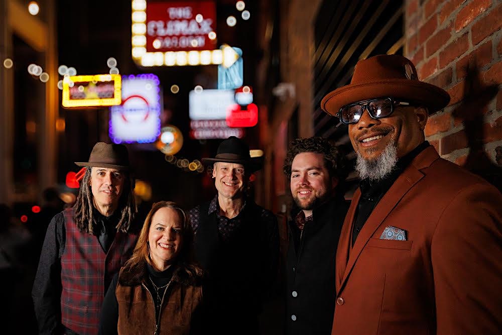 Five musicians pose on a city sidewalk at night, lit by colorful neon signs from nearby venues.