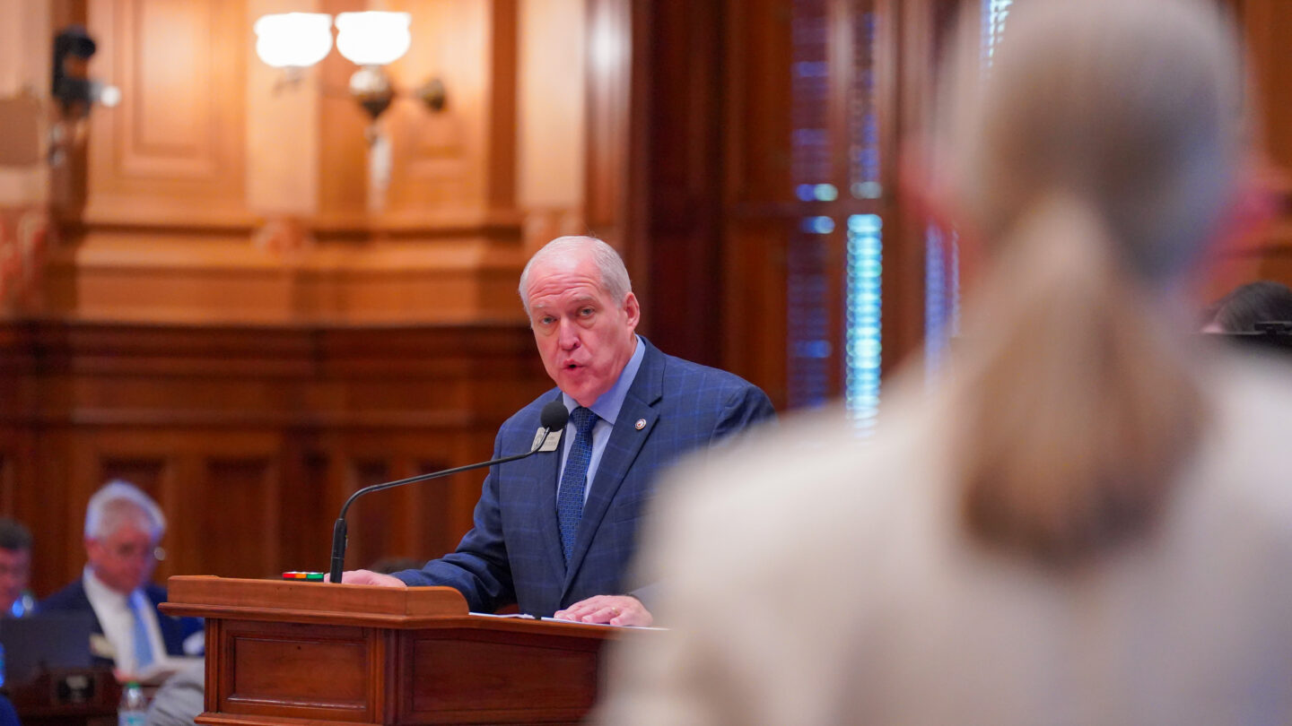 A lawmaker speaking behind a podium looks toward a standing person.