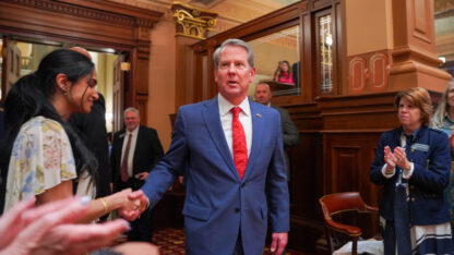 Brian Kemp, wearing a blue suit and a red tie, walks into the House chamber surrounding by people applauding him