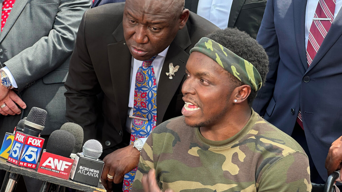A man sitting in a lchair speaks into several microphones at a press conference while surrounded by people