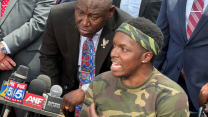 A man sitting in a lchair speaks into several microphones at a press conference while surrounded by people