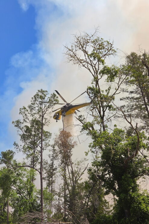 A helicopter sprays a substance to fight an ongoing fire as smoke rises into the air above some trees