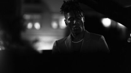 Jon Batiste at a piano in closeup, eyes closed, stage lights visible in the background, black and white photograph.