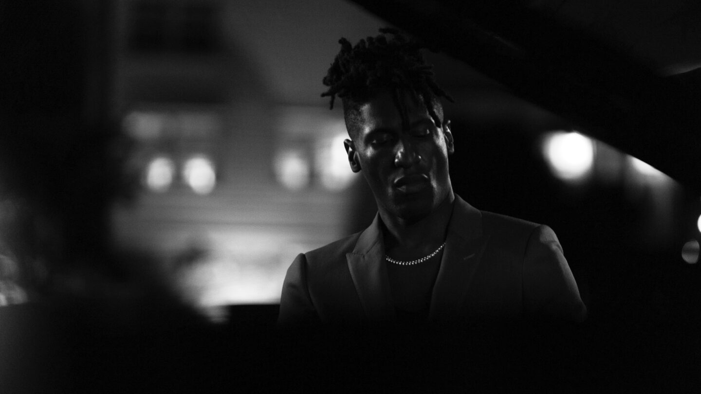Jon Batiste at a piano in closeup, eyes closed, stage lights visible in the background, black and white photograph.