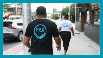 The back of a person wearing a black shirt that says PAD in blue letters and a person wearing a white shirt that says PAD in blue letters.