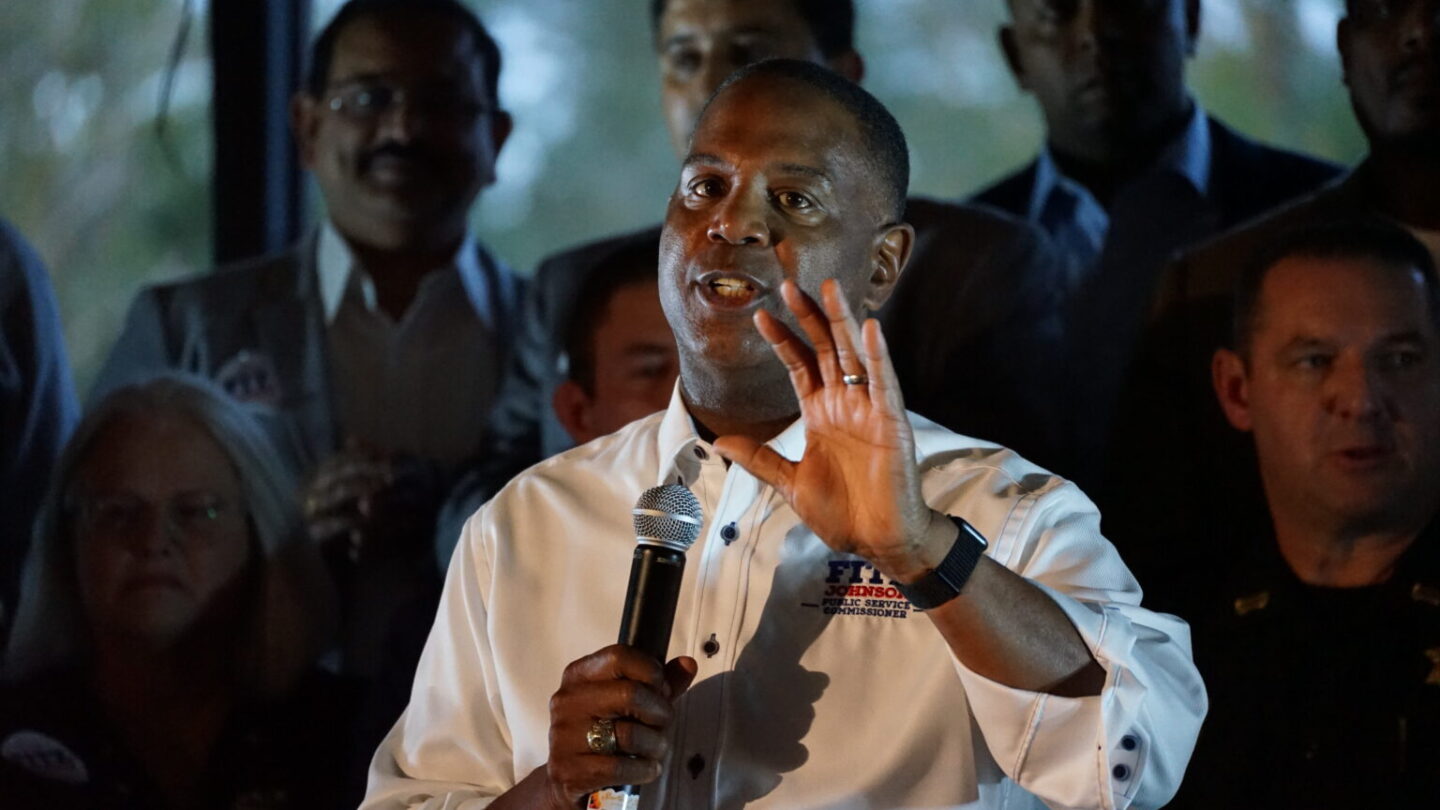 A man stands holding his left hand out while holding a microphone with his right hand. A group of people stand behind him