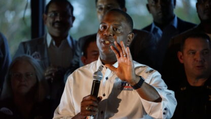 A man stands holding his left hand out while holding a microphone with his right hand. A group of people stand behind him