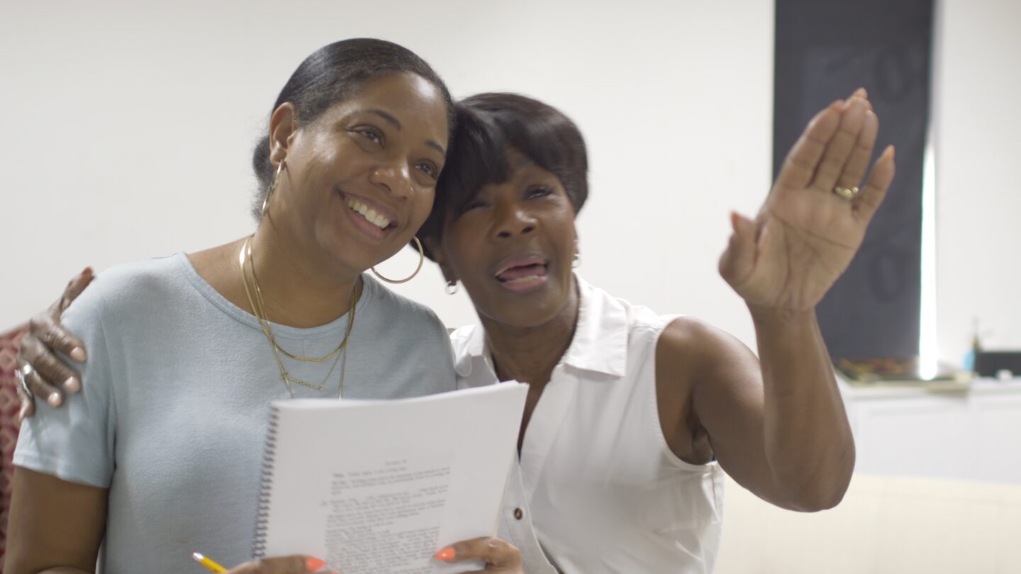 Two women laugh together during a rehearsal, one holding a spiral-bound script