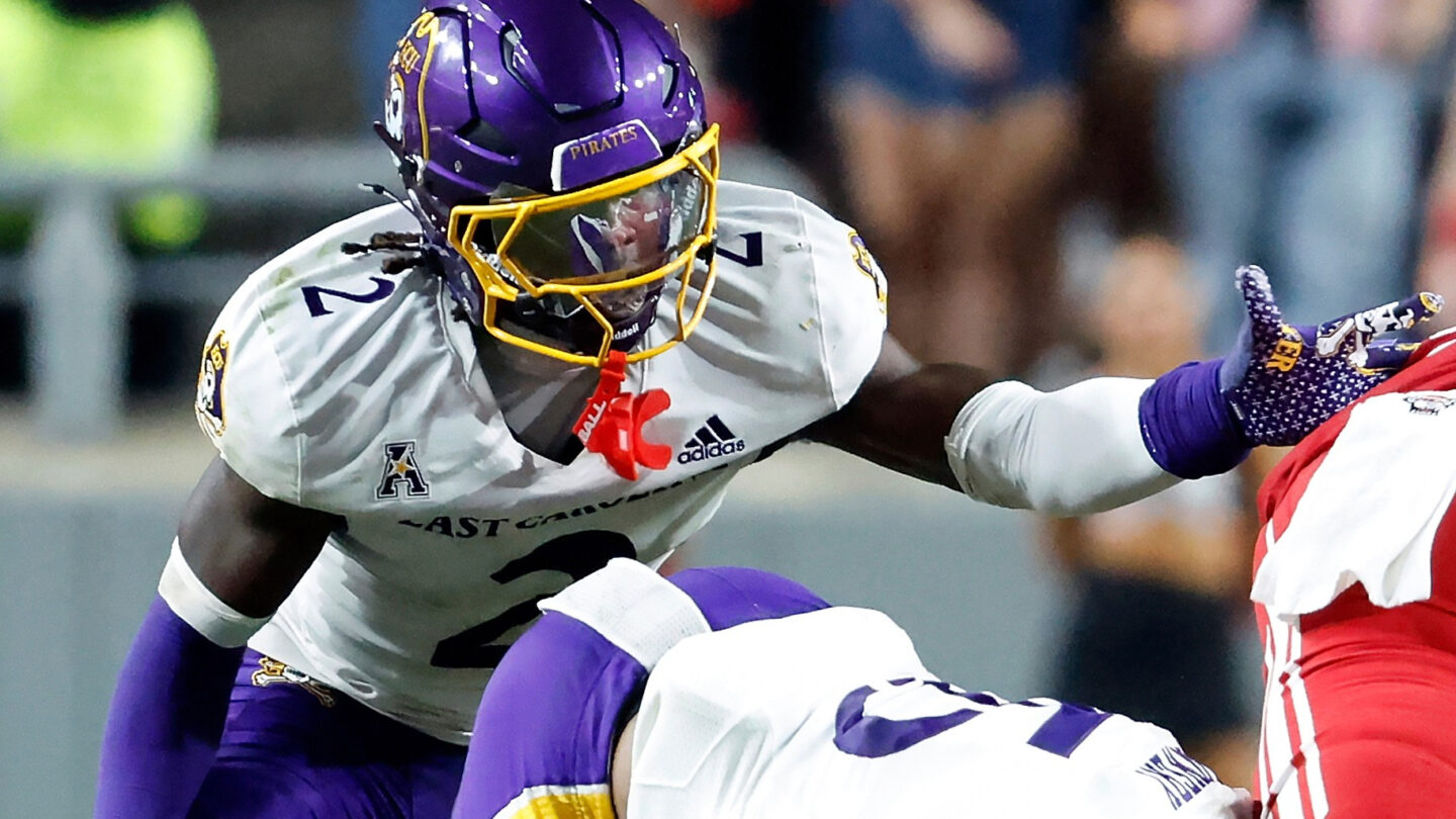 A football player in a purple helmet and a white and purple uniform helps with a tackle
