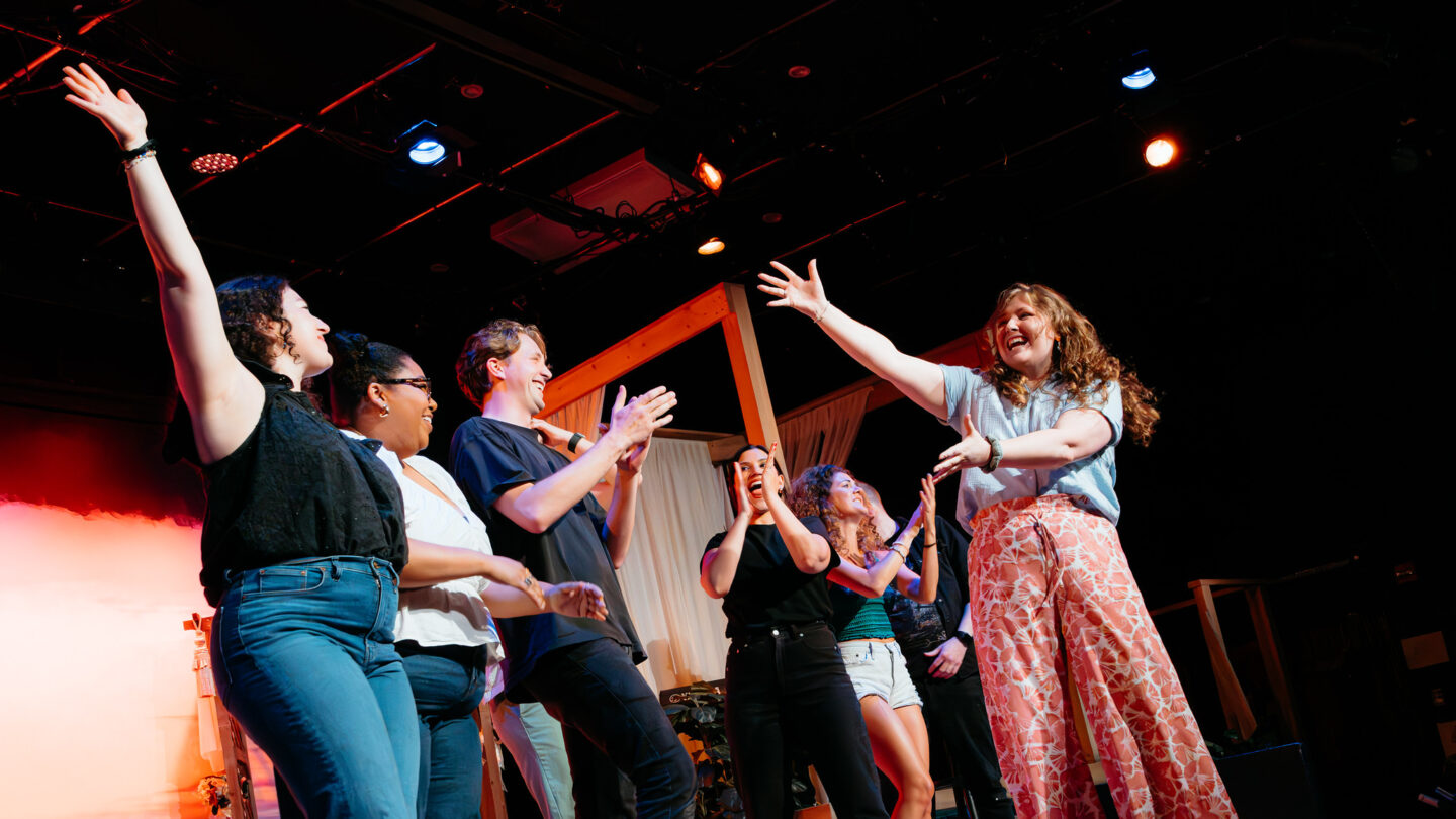 Cast of Road Trip 2 takes a celebratory bow on stage at the 2026 Atlanta Fringe Festival, arms raised and clapping under stage lighting.