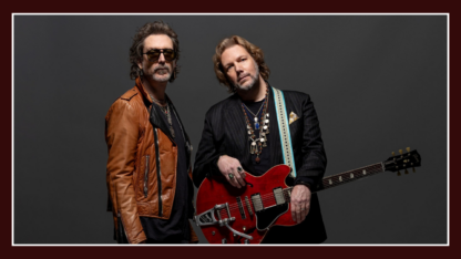 Chris and RIch Robinson pose in front of a dark grey background. One holds a guitar.