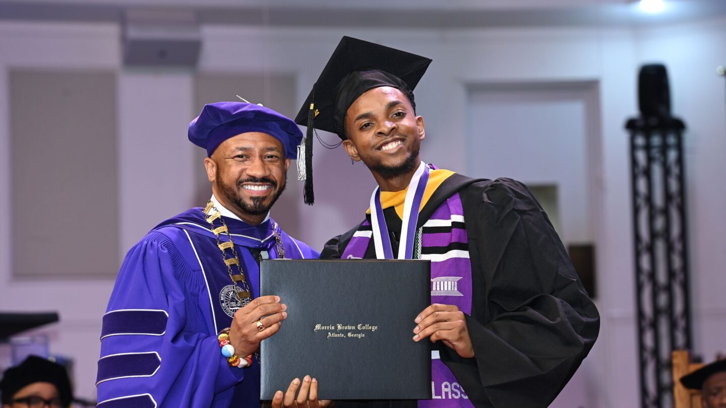 Morris Brown College graduate Shandell-Bright Johnson in a black cap and gown with sash poses with Morris Brown President Dr. Kevin James in a purple cap and gown holding a diploma.