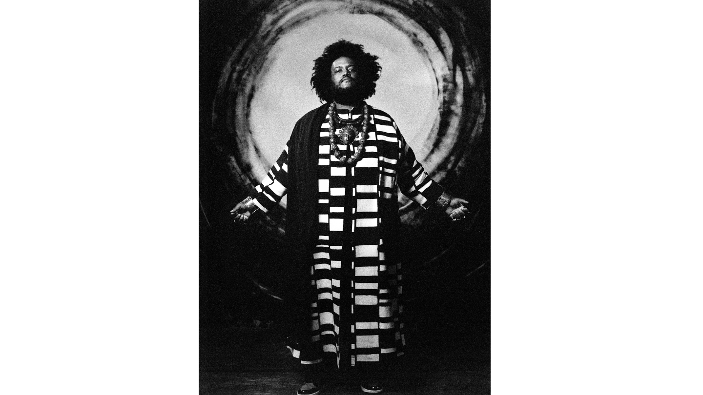 Kamasi Washington stands in bold black-and-white striped robes against a circular backdrop.