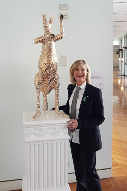 Olivia Newton-John stands beside a gold mosaic kangaroo sculpture in a gallery setting, smiling at the camera.