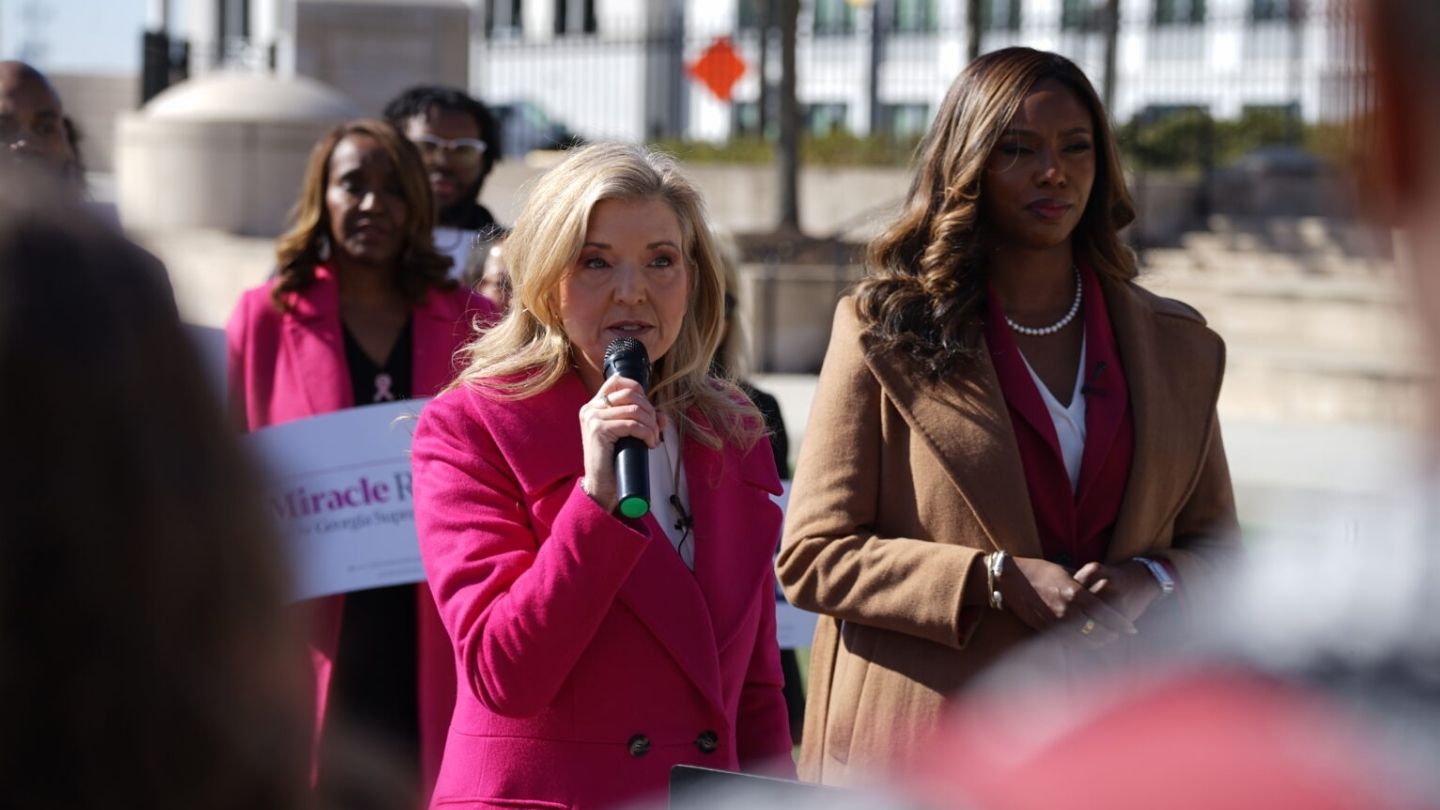 Two women stand while one in a pink suit talks into a microphone