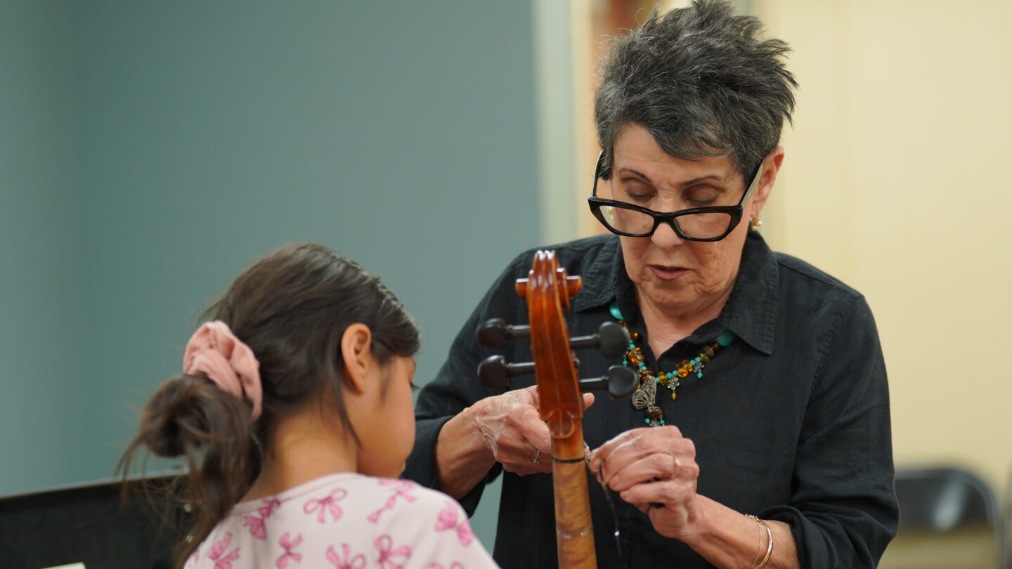 A woman tapes lines on the neck of a cello while a child stands in front of her