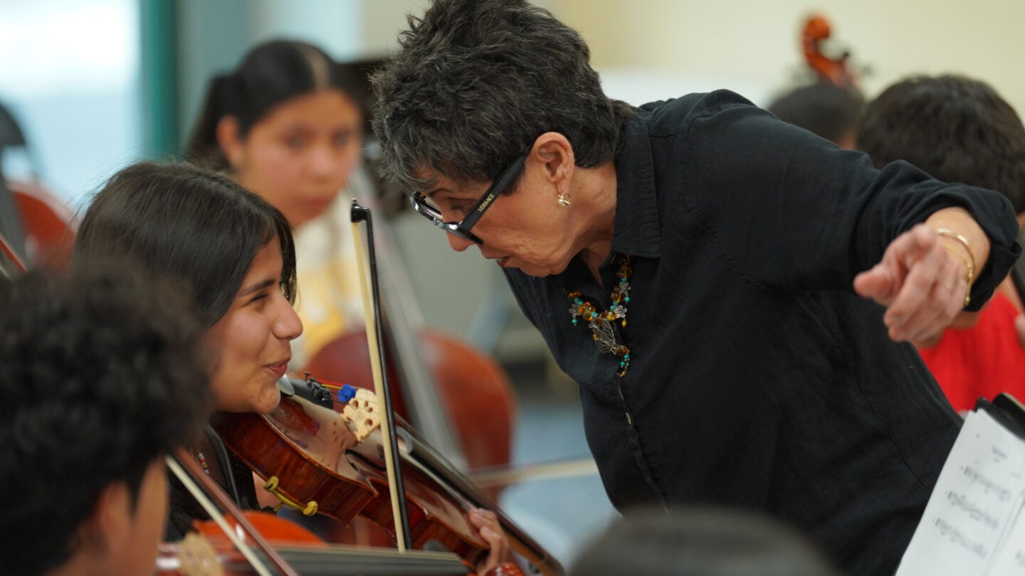 A woman leans forward while several students play stringed instruments