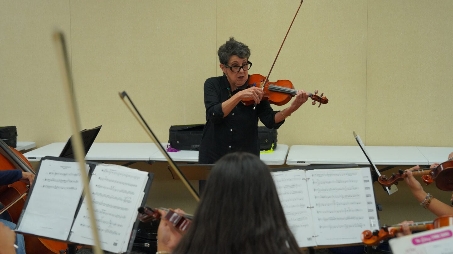 A conductor standing at the head of an orchestra plays a violin