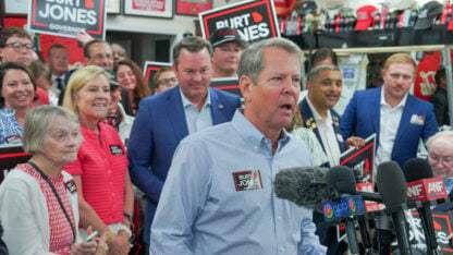 Brian Kemp speaks into several microphones at an indoor campaign event surrounded by people