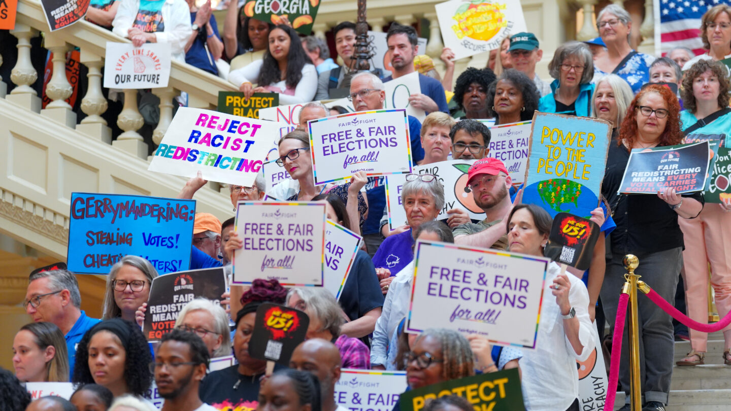 A large group of protesters stand on white marble stairs, holding a myriad of colorful signs