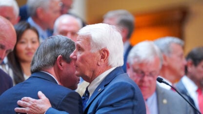 A man speaks into the ear of another man, while other lawmakers stand behimd them