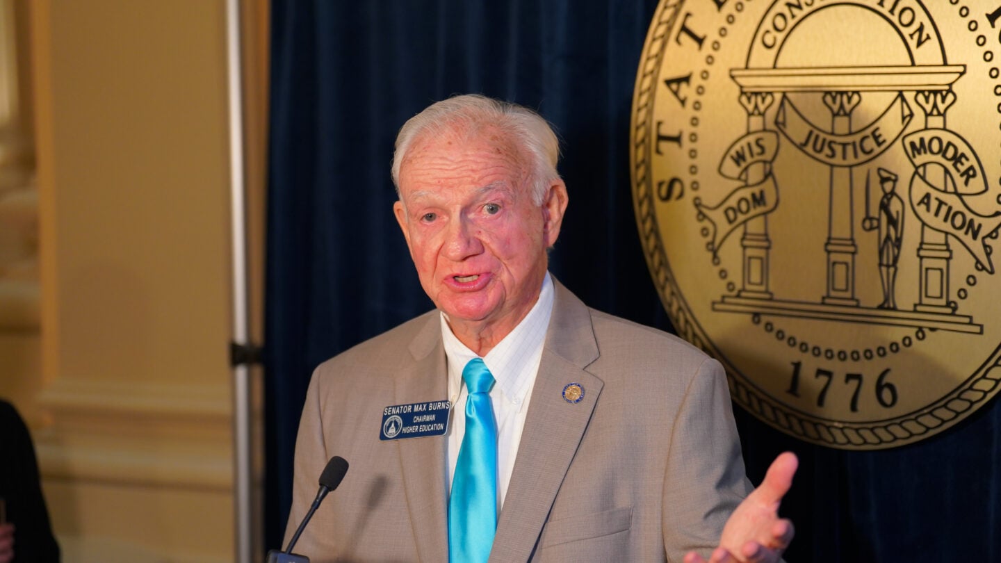A man stands in front of a gold seal and speaks into a microphone