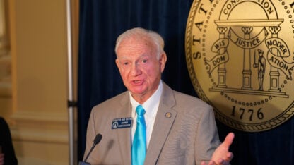 A man stands in front of a gold seal and speaks into a microphone