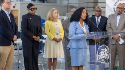 A group of people stand behind a woman wearing a light blue blazer and skirt who is speaking at a podium indoors