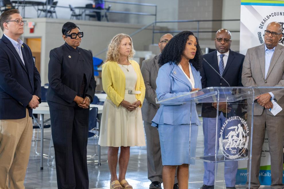 A group of people stand behind a woman wearing a light blue blazer and skirt who is speaking at a podium indoors