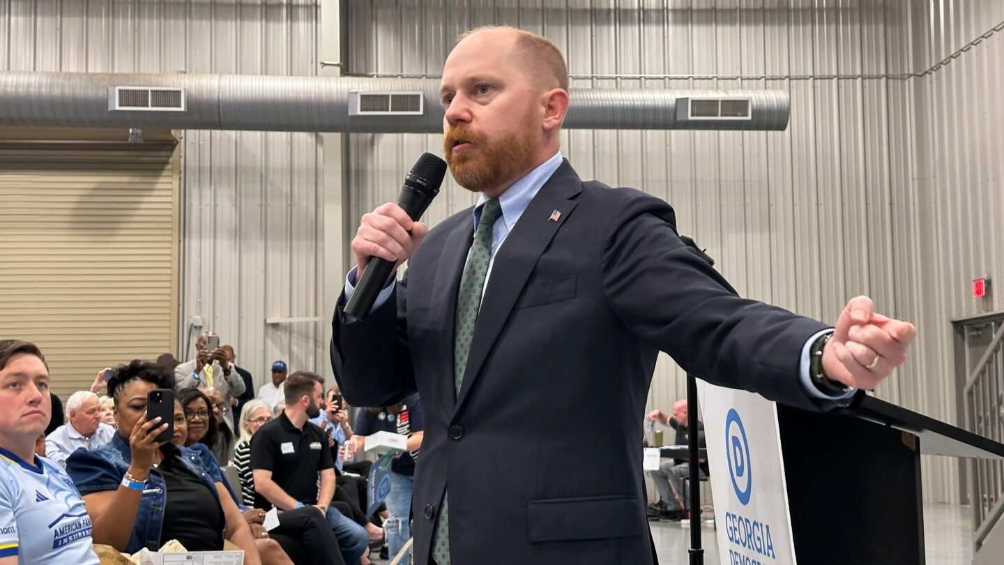 A man in a suit speaks into a microphone in front of a crowd