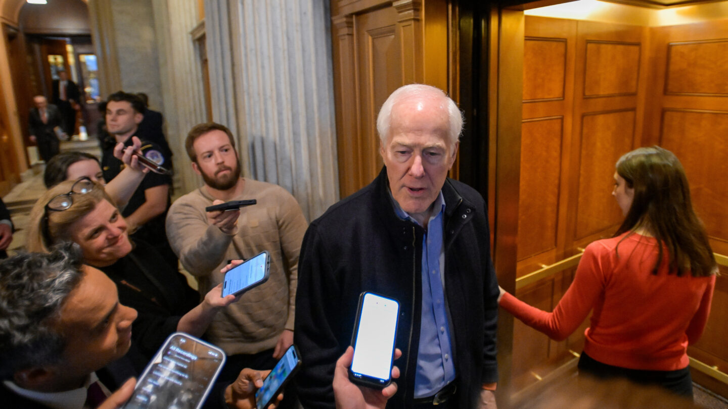 A man with grey hair in a blue button-down shirt answers questions from reporters before boarding an elevator