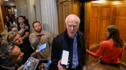 A man with grey hair in a blue button-down shirt answers questions from reporters before boarding an elevator
