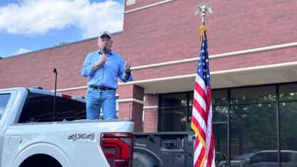 A man in jeans and a button-down shirt stands in the bed of a pickup truck with a microphone in his hand and an american flag on his right