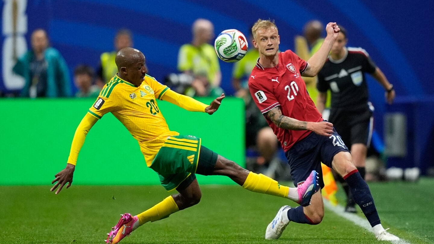 A soccer player extends his right leg to kick a ball while another player runs alongside him