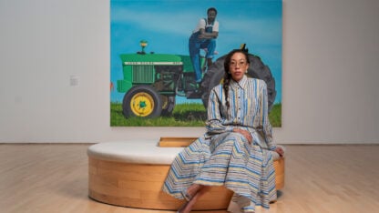 Amy Sherald, wearing glasses and a striped dress, sits on a circular wooden bench in front of her large-scale painting of a Black man on a green John Deere tractor.