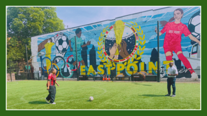 Two people kick a soccer ball in front of a large mural depicting soccer players