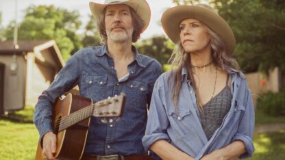 Gillian Welch and David Rawlings, headliners of the 2026 Amplify Decatur Music Festival, stand outdoors. Rawlings holds an acoustic guitar. Both wear wide-brim hats.