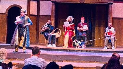 Five actors in a mix of period costumes and street clothes perform on the Shakespeare Tavern Playhouse stage, prompt scripts in hand, with a small dog on a leash visible center stage alongside the ensemble.