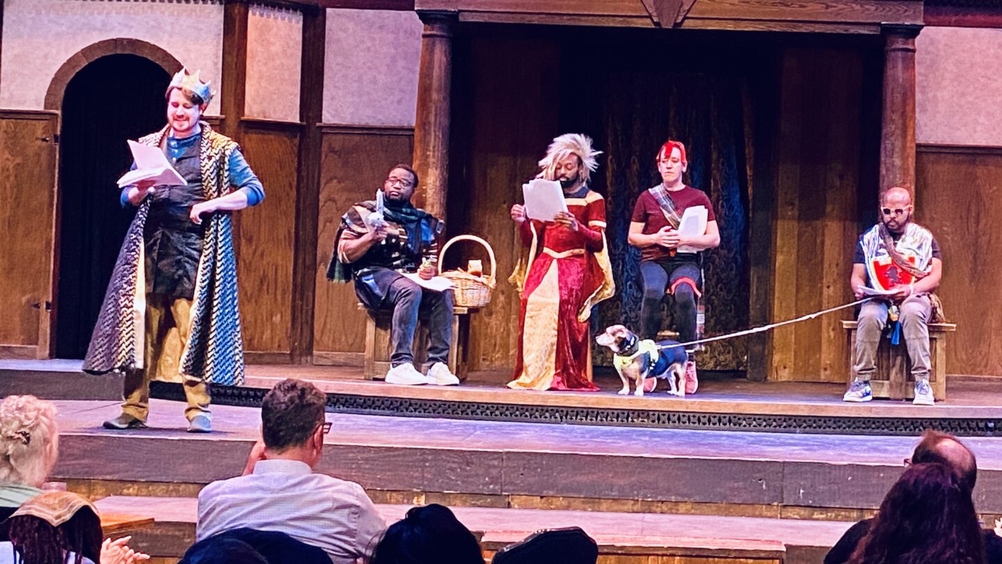 Five actors in a mix of period costumes and street clothes perform on the Shakespeare Tavern Playhouse stage, prompt scripts in hand, with a small dog on a leash visible center stage alongside the ensemble.