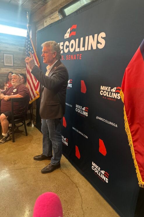 Mike Collins speaks and stands in front of a backdrop that has his name on it