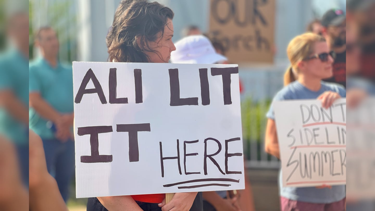 A person holds up a sign that says Ali Lit It Here.