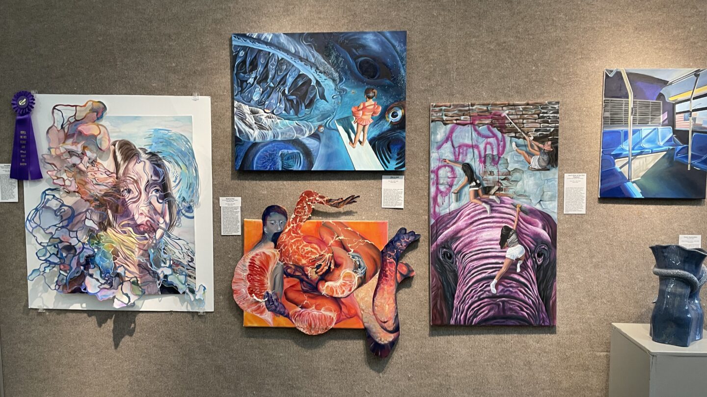 Five mixed-media and painted works hang on a gray exhibition wall, ranging from a flowing figurative piece with a blue ribbon at left to a large painting of a girl climbing a purple elephant at center right.