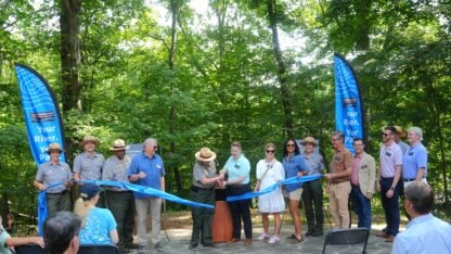 A group of people cut a blue ribbon in front of a podium in the forest.
